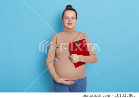 Good looking caucasian pregnant lady holding floor scales in hand and touch her belly, looks smiling at camera, being glad of result of measurement, isolated over blue background. 101323863
