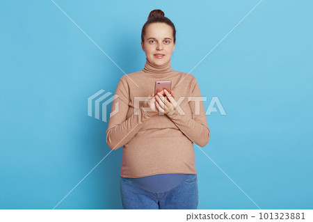 European pregnant with with pleasant appearance, holding phone in hands and looking directly at camera, needs to make call, wearing casually, isolated over blue background. 101323881
