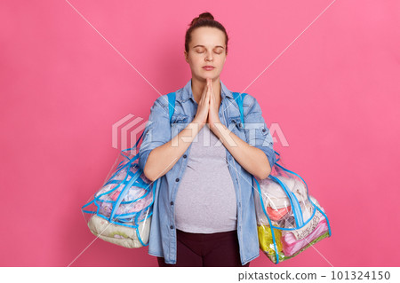 Beautiful pregnant woman with hair bun,expecting baby, keeps palms together and prays, posing isolated on pink background, wearing casual clothing, holding bags with stuff for maternity house. Beautiful pregnant woman with hair bun,expecting baby, keeps palms together and prays, posing isolated on pink background, wearing casual clothing, holding bags with stuff for maternity house. 101324150