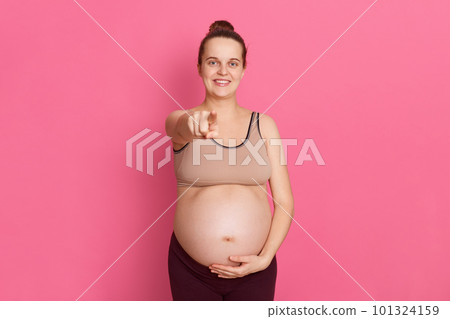 Young pregnant woman pointing finger to camera and touching her bare belly with other hand, looks happy, has hair bun, dresses leggins and tank top, standing isolated over pink background. Young pregnant woman pointing finger to camera and touching her bare belly with other hand, looks happy, has hair bun, dresses leggins and tank top, standing isolated over pink background. 101324159