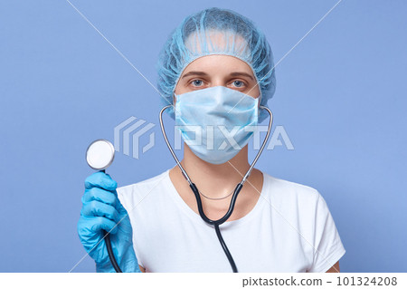 Horizontal shot of doctor woman holding stethoscope in front of her, image isolated over blue background, wearing white t shirt, disposable hat, gloves and protective mask. Medicine concept. Horizontal shot of doctor woman holding stethoscope in front of her, image isolated over blue background, wearing white t shirt, disposable hat, gloves and protective mask. Medicine concept. 101324208