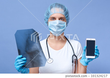 Indoor shot of female doctor holding x ray scan and being ready to talk on phone with diagnostic center, posing isolated over blue studio background, looking at camera, has stethoscope. Copy space. Indoor shot of female doctor holding x ray scan and being ready to talk on phone with diagnostic center, posing isolated over blue studio background, looking at camera, has stethoscope. Copy space. 101324217