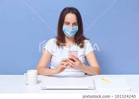 Woman dresses white casual t shirt and protective mask holding phone in hands, sees some important news about disease, has surprise and shock in eyes. Young girl with dark hair sitting at white table. Woman dresses white casual t shirt and protective mask holding phone in hands, sees some important news about disease, has surprise and shock in eyes. Young girl with dark hair sitting at white table. 101324240