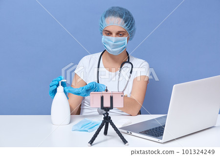 Close up portrait of medical doctor works online from home, helping people in days of panic and pandemic, shows in her vlog how to use sanitaizer, sitting at white desk in front of tripod with phone. 101324249
