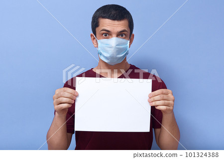 Indoor picture of surprised handsome young man holding sheet of paper in both hands, space for text, wearing antibacterial mask, conveying message, being in quarantine. Covid19 outbreak concept. 101324388