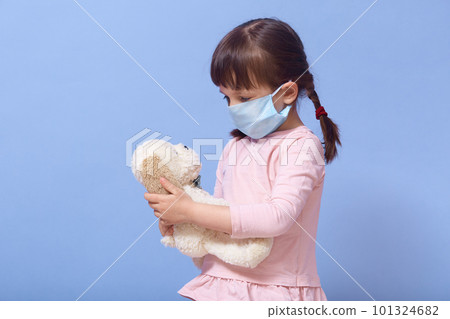 Close up portrait of little girl wearing casual clothing and medical mask, holding puppy toy in hands and looking at it, female child with pigtails isolated over blue studio background. Coronavirus. Close up portrait of little girl wearing casual clothing and medical mask, holding puppy toy in hands and looking at it, female child with pigtails isolated over blue studio background. Coronavirus. 101324682