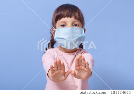 Closeup portrait of cute child wearing casual shirt and medical mask, female kid showing stop gesture with both palms, looking at camera, has two pigtails, posing isolated over blue studio background. 101324693