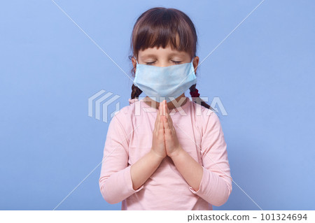 Coronavirus, self isolation, quarantine concept. Little Caucasian girl wearing mask for protect fron covid 19, standing with closed eyes and praying for stop corona virus, poses against blue wall. 101324694