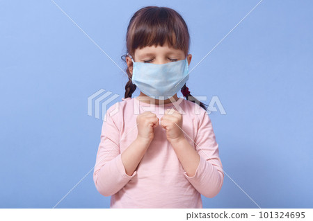 Image of upset sad little girl with pigtails closing her eyes, keeping both fists, praying, being scared with coronavirus, wearing antibacterial mask, being alone. Children and covid 19 concept. Image of upset sad little girl with pigtails closing her eyes, keeping both fists, praying, being scared with coronavirus, wearing antibacterial mask, being alone. Children and covid 19 concept. 101324695