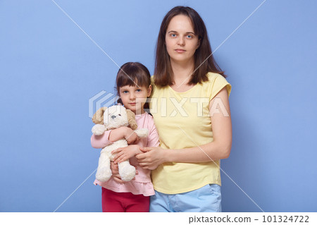Indoor shot of beautiful blue eyed female hugging her daughter, little girl holding her favourite toy, looking directly at camera, family spending time together, being in good mood. Family concept. 101324722