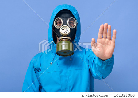Studio shot of man dresses chemical protective suit and respirator posing and showing stop gesture with his hand against blue wall. Passage is forbidden. Air pollution, danger of infection concept. 101325096