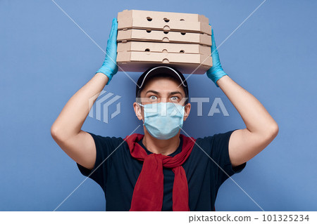 Studio shot of surprised delivery man dresses casual outfit, mask and latex gloves, holding stack of pizza's boxes above his head, has big eyes, looks shocked, delivers online order to customer. 101325234