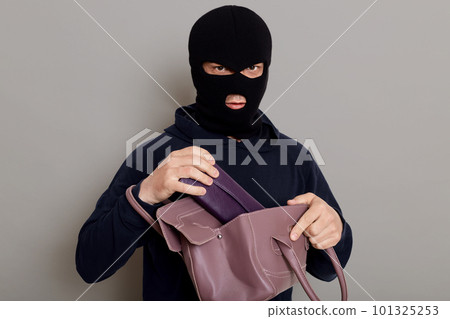 A criminal thief takes a wallet from a stolen bag, dressed in a black hoodie and a robbery mask, looking directly at camera, isolated on gray background. A criminal thief takes a wallet from a stolen bag, dressed in a black hoodie and a robbery mask, looking directly at camera, isolated on gray background. 101325253
