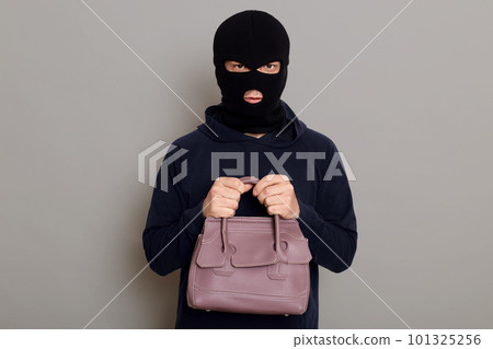 A young guy thief is dressed in a black hoodie and robbery mask, holds a woman's bag with both hands, stole other people's things, looks at the camera, isolated on gray background. 101325256