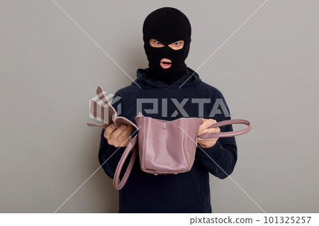 Young male criminal with stolen bag in his hands, looks in inside with shocked expression on his face, being very surprised that he sees in bag, isolated over gray background. 101325257