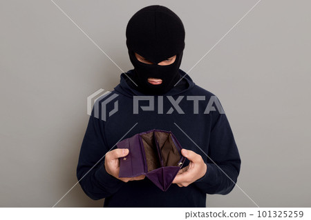 Frustrated guy thief dressed in a black hoodie and a robbery mask, holding a stolen open empty wallet in his hands, looks upset, isolated on gray background. 101325259