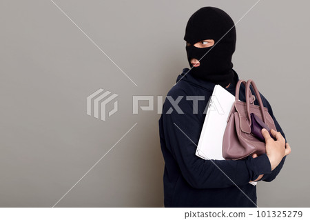 A young thief male dressed in a black hoodie with a disguised face runs away after stealing with other people's belongings, bag, laptop, wallet. Look around, be afraid of being caught, copy-space. A young thief male dressed in a black hoodie with a disguised face runs away after stealing with other people's belongings, bag, laptop, wallet. Look around, be afraid of being caught, copy-space. 101325279