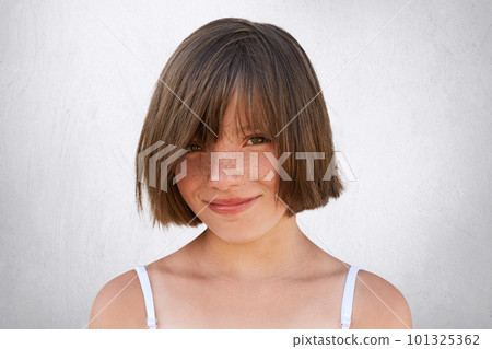 Smiling little girl with stylish hairdo, dark eyes and freckled face posing against white background. Pretty girl with happy expression looking at camera with eyes full of happiness. Child concept 101325362