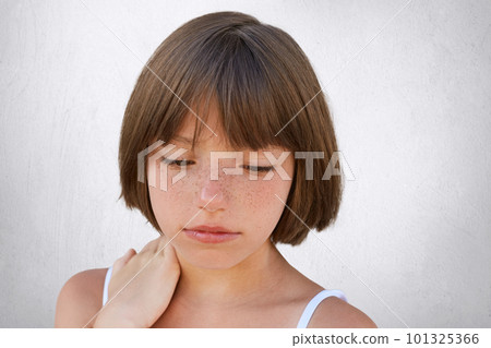 Close up of attractive little child with freckles and dark short hair keeping her hand on neck, looking seriously down, having thoughtful expression while posing against white concerete wall 101325366
