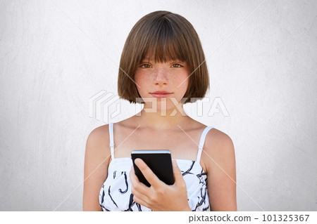 Childhood in digital age. Cute girl with short stylish hairdo, dark deep-set eyes and freckles wearing nice dress, holding smart phone in her hands, playing games online isolated on white wall Childhood in digital age. Cute girl with short stylish hairdo, dark deep-set eyes and freckles wearing nice dress, holding smart phone in her hands, playing games online isolated on white wall 101325367