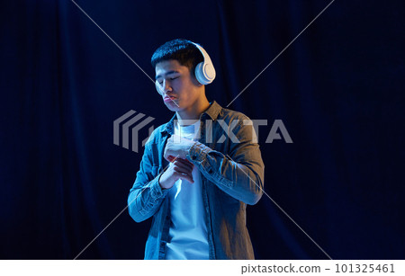 Evaluating songs. Young asian guy in casual stylish clothes listening to music in headphones against dark background in neon light. Concept of human emotions and lifestyle 101325461