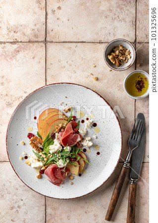 Salad of arugula and prosciutto or ham with the addition of sliced apple, cheese, walnuts and spices on a plate with cutlery on pink tile background. Top view, text space. Salad of arugula and prosciutto or ham with the addition of sliced apple, cheese, walnuts and spices on a plate with cutlery on pink tile background. Top view, text space. 101325696