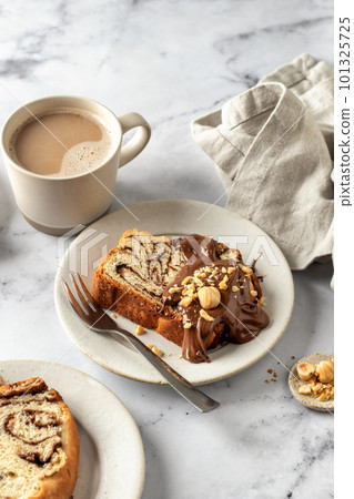 Slice of brioche babka bread with chocolate filling on white marble background served with chocolate and nuts, coffee cup. Easter traditional baking Slice of brioche babka bread with chocolate filling on white marble background served with chocolate and nuts, coffee cup. Easter traditional baking 101325725