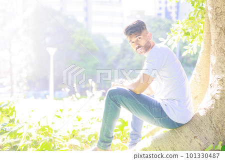 An Indian man sitting under a tree in a park with sunlight filtering through the trees 101330487