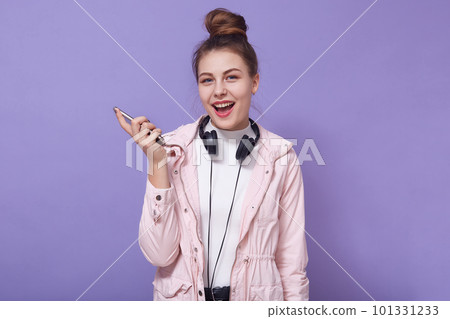 Happy excited woman wearing casual clothing,having headphones around neck, holding modern smart phone in hands, posing with opened mouth against lilac wall, adorable lady enjoying listening to music. Happy excited woman wearing casual clothing,having headphones around neck, holding modern smart phone in hands, posing with opened mouth against lilac wall, adorable lady enjoying listening to music. 101331233