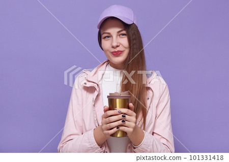 Close up portrait of charismatic charming attractive young female holding themo mug in both hands, drinking coffee, having walk, wearing pink cap and leather jacket, white sweater. Youth concept. 101331418