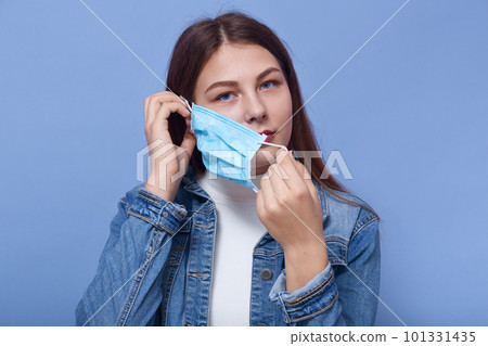 Close up portrai tof attractive female puts on or takes off her mask isolated over blue background, lady wearing white shirt and denim jacket, looking directly at camera.Coronavirus concept. Close up portrai tof attractive female puts on or takes off her mask isolated over blue background, lady wearing white shirt and denim jacket, looking directly at camera.Coronavirus concept. 101331435