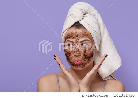 Attractive lady making funny face while doing peeling procedures for face at home, keeping palms near cheeks, looking aside, has white towel on head, keeps lips rounded. Attractive lady making funny face while doing peeling procedures for face at home, keeping palms near cheeks, looking aside, has white towel on head, keeps lips rounded. 101331436