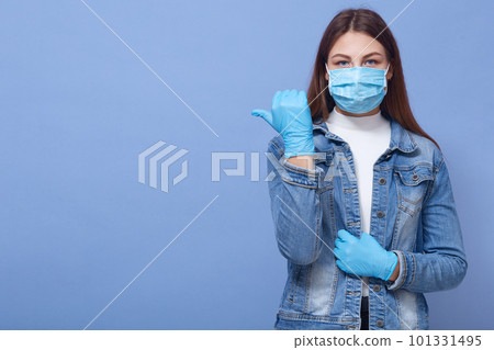 Indoor photo of confident attractive young woman looking directly at camera, making gesture, showing direction with thumb, wearing gloves and antibacterial mask. Copyspace for advertisement. Indoor photo of confident attractive young woman looking directly at camera, making gesture, showing direction with thumb, wearing gloves and antibacterial mask. Copyspace for advertisement. 101331495
