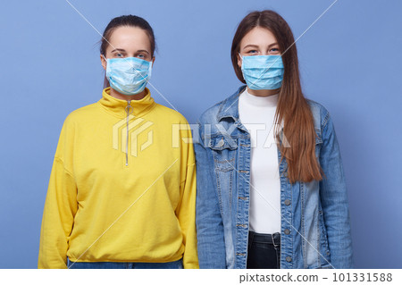 Close up portrait of two women wearing casual outfit and health masks, trying to prevent from flu, coronavirus or contagious diseases, posing isolated over blue background, females looking at camera. 101331588