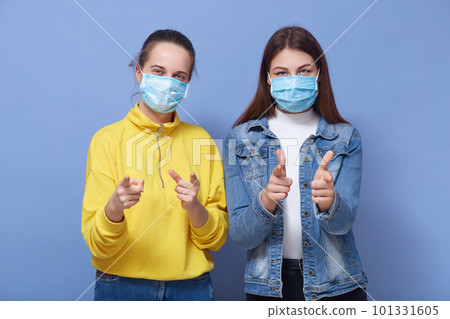Indoor studio picture of two charismatic good looking females wearing protection masks, making gestures, showing direction with forefingers, standing isolated over blue background. Covid 19 concept. Indoor studio picture of two charismatic good looking females wearing protection masks, making gestures, showing direction with forefingers, standing isolated over blue background. Covid 19 concept. 101331605