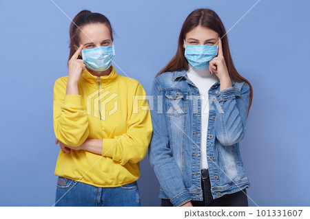 Two young women wearing protection mask for coronavirus disease over blue background thinking concentrated about doubt with finger on temples and looking at camera wondering. Two young women wearing protection mask for coronavirus disease over blue background thinking concentrated about doubt with finger on temples and looking at camera wondering. 101331607