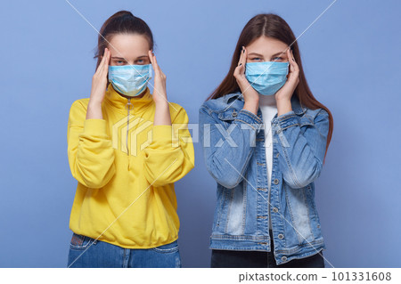 Portrait of two young dark haired girls wearing flu medicine mask and casual attires, standing isolated over background, keeping hands on head, suffering from headache. Portrait of two young dark haired girls wearing flu medicine mask and casual attires, standing isolated over background, keeping hands on head, suffering from headache. 101331608