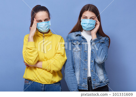Studio portrait of two young girls with headache, holding hands on temples, wearing medical flu mask and posing iolated over blue background, females trying to avoid coronavirus. Covid 19 concept. Studio portrait of two young girls with headache, holding hands on temples, wearing medical flu mask and posing iolated over blue background, females trying to avoid coronavirus. Covid 19 concept. 101331609