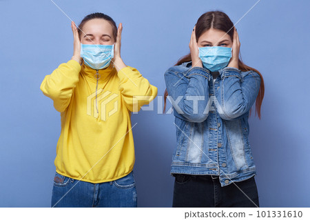 Horizontal shot of young Caucasian women wearing casual clothing covering ears and closed eyes, ladies with medical disposable mask, females protective from coronavirus. Covid 19, healthcare concept. Horizontal shot of young Caucasian women wearing casual clothing covering ears and closed eyes, ladies with medical disposable mask, females protective from coronavirus. Covid 19, healthcare concept. 101331610