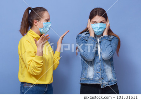 Horizontal picture of one young female with ponytail raising hands, shouting at her friend, being in isolation, wearing masks, being in quarantine, having quarrels. People and communication concept. Horizontal picture of one young female with ponytail raising hands, shouting at her friend, being in isolation, wearing masks, being in quarantine, having quarrels. People and communication concept. 101331611