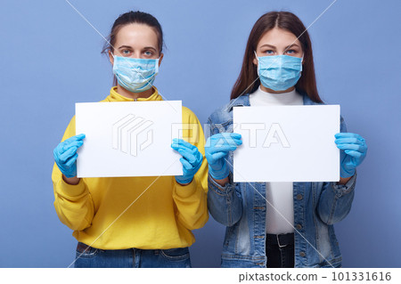Image of two young women wearing medical disposable masks and medical gloves holding white blank papers in hands, copy space for some inscriptions. Corona virus, covid 19, health care concept. Image of two young women wearing medical disposable masks and medical gloves holding white blank papers in hands, copy space for some inscriptions. Corona virus, covid 19, health care concept. 101331616