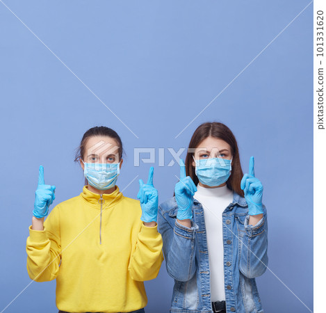 Pretty women wearing medical masks and latex gloves pointing index fingers isolated over blue background, copy space fore advertisement or promotional text. Pretty women wearing medical masks and latex gloves pointing index fingers isolated over blue background, copy space fore advertisement or promotional text. 101331620