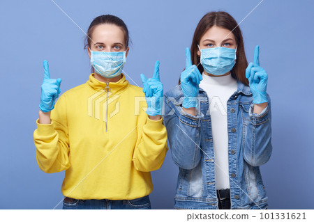 Covid 19, coronavirus concept. Young Caucasian women wearing medical mask and gloves, casual outfits, standing and pointing up with index fingers, beautiful ladies trying to avoid virus infection. Covid 19, coronavirus concept. Young Caucasian women wearing medical mask and gloves, casual outfits, standing and pointing up with index fingers, beautiful ladies trying to avoid virus infection. 101331621