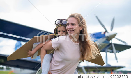 Beautiful caucasian woman is holding her little daughter at the airfield. A small girl in a pilot's costume sits on her mother's back against the background of the plane. 101332553