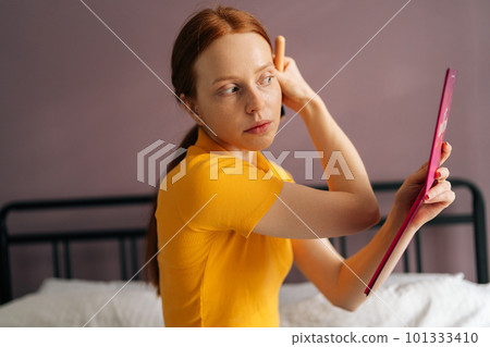 Focused redhead young woman with natural beauty applying powder or blusher on face with big makeup brush sitting on bed, looking to mirror. Pretty female putting daily makeup putting cosmetic on face. Focused redhead young woman with natural beauty applying powder or blusher on face with big makeup brush sitting on bed, looking to mirror. Pretty female putting daily makeup putting cosmetic on face. 101333410