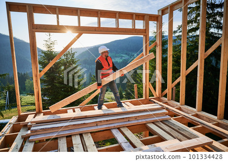 Carpenter constructing wooden-framed house near the woods at sunset. Man holding large joist and dressed in work clothes and helmet. Concept of modern and environmentally-friendly construction. 101334428