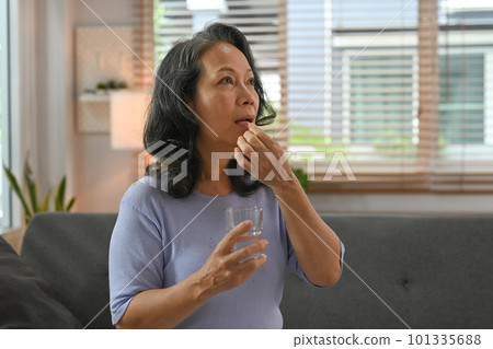 Middle aged woman sitting on couch holding pill and glass of fresh water. Pharmaceutical healthcare treatment concept 101335688