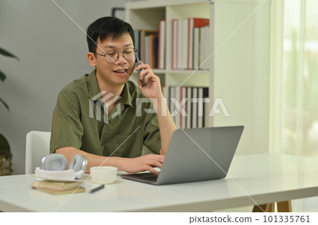 Positive millennial man in glasses talking on cellphone, sitting at working desk with laptop computer 101335761