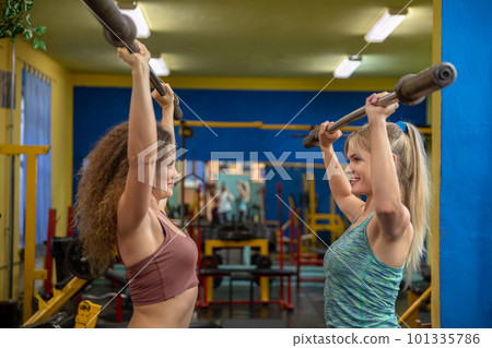 Two women at the gym lift barbells over their heads 101335786