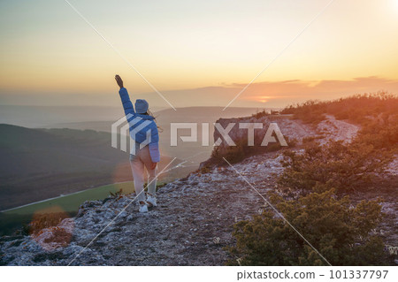 Woman hiker open arms on top of sunrise mountain. The girl salutes the sun, wearing a blue jacket, white hat and white jeans. Conceptual design. 101337797